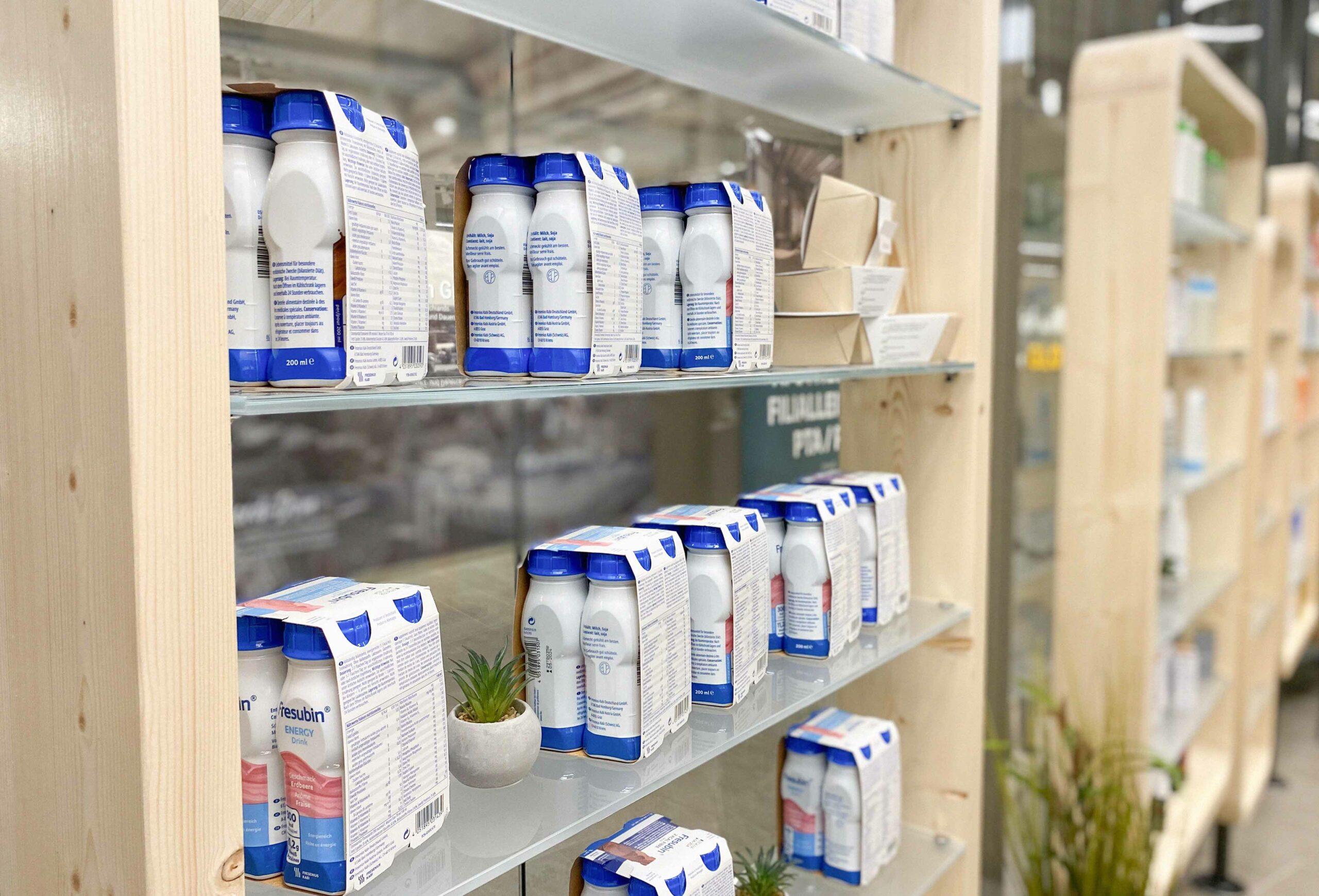 Nahaufnahme eines Regals in der Wesel Apotheke der Dr. Kottenberg Apotheken. Zu sehen sind mehrere Packungen medizinischer Trinknahrung der Marke Fresubin in weißen Flaschen mit blauem Deckel. Die Produkte sind ordentlich auf Glasböden in einem hellen Holzregal platziert.