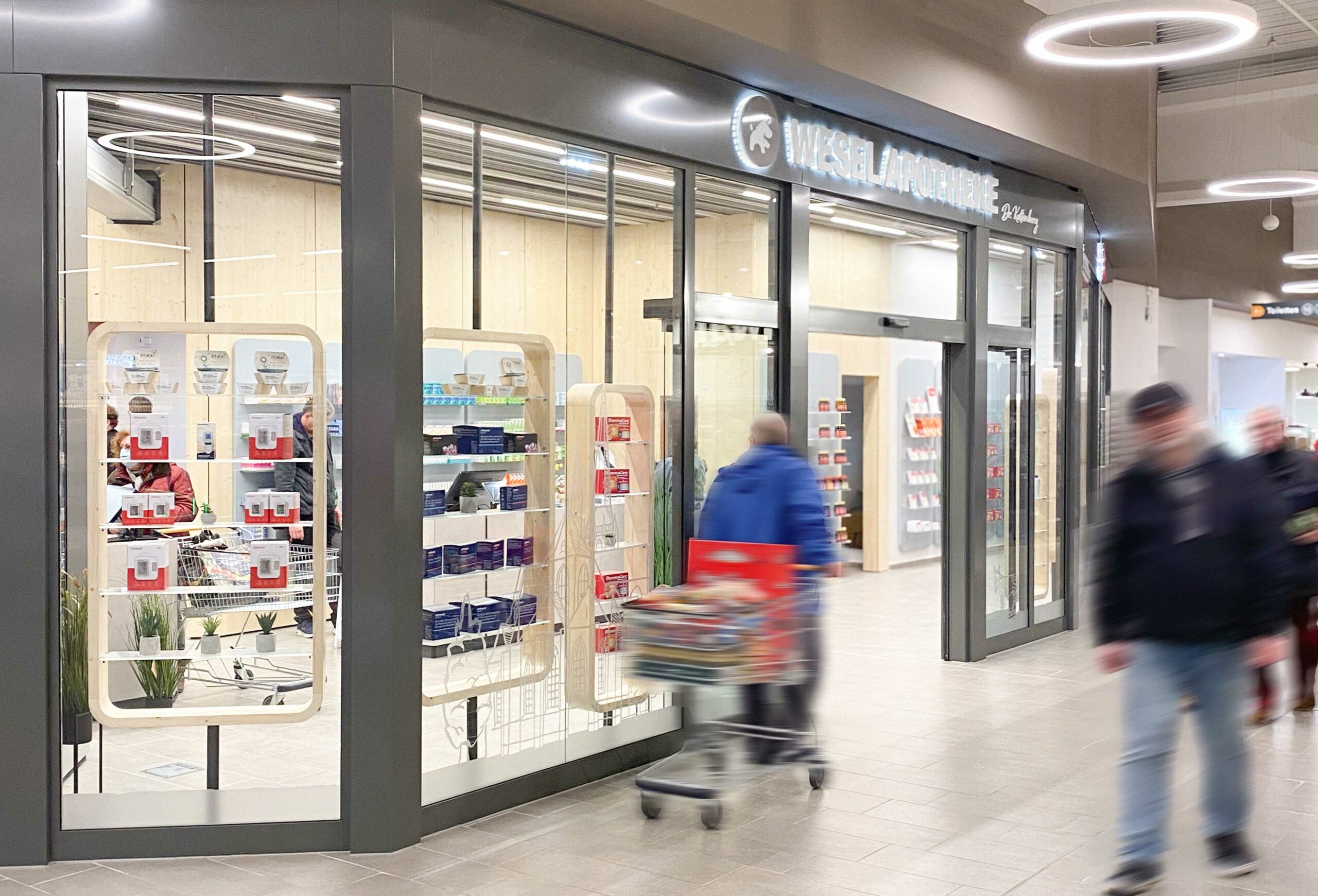 Die moderne Eingangsfassade der Wesel Apotheke im Einkaufszentrum mit großen Glasfenstern und hell beleuchtetem Innenraum. Im Vordergrund gehen mehrere Menschen mit Einkaufswagen vorbei.
