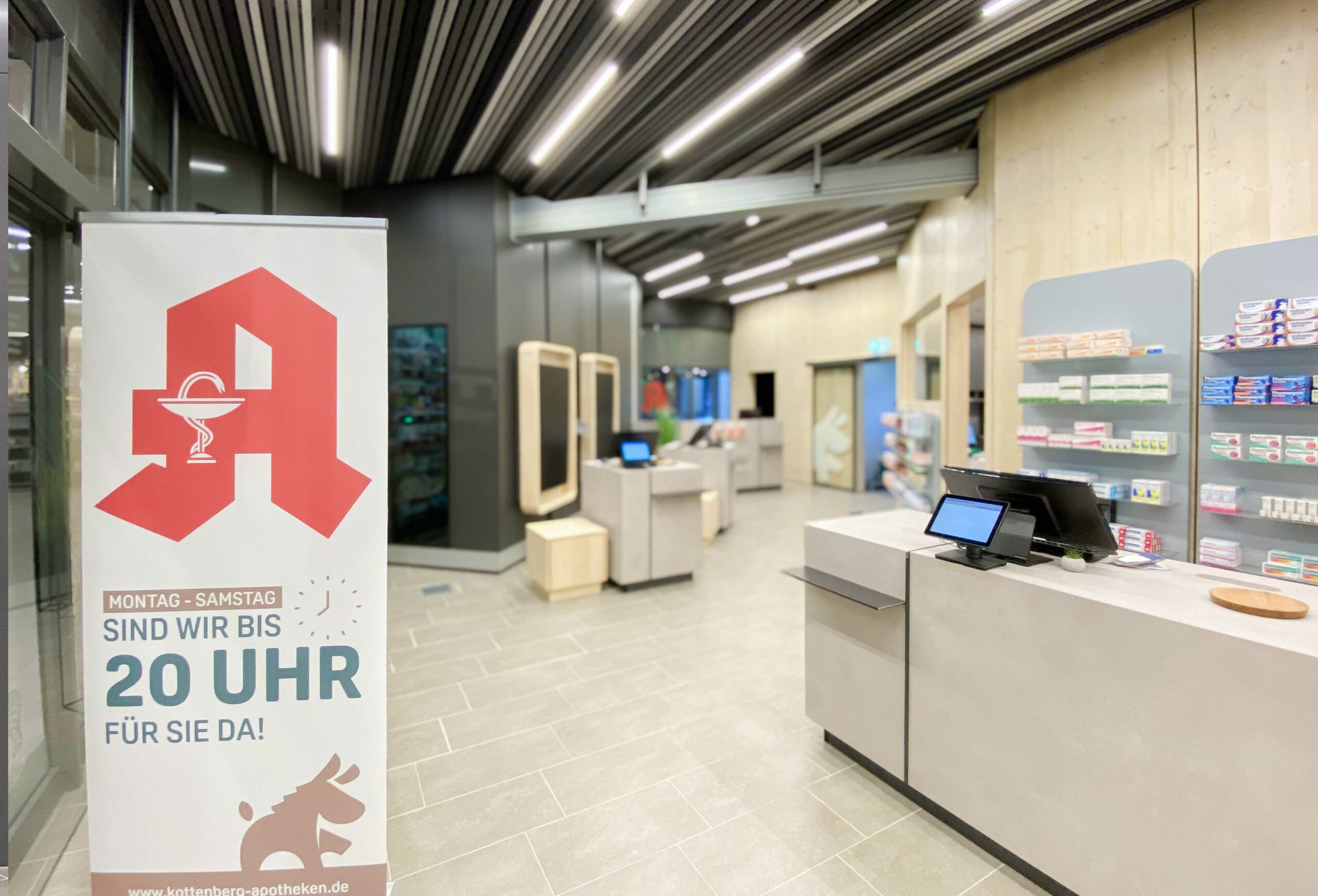 Innenansicht der Wesel Apotheke der Dr. Kottenberg Apotheken. Im Vordergrund ein modernes Verkaufsterminal, im Hintergrund ein aufgestellter Banner mit dem Apotheken-A und dem Hinweis: „Montag – Samstag sind wir bis 20 Uhr für Sie da!“. Die Apotheke ist hell, offen und zeitgemäß gestaltet, mit digitaler Ausstattung und klar strukturierten Regalen.