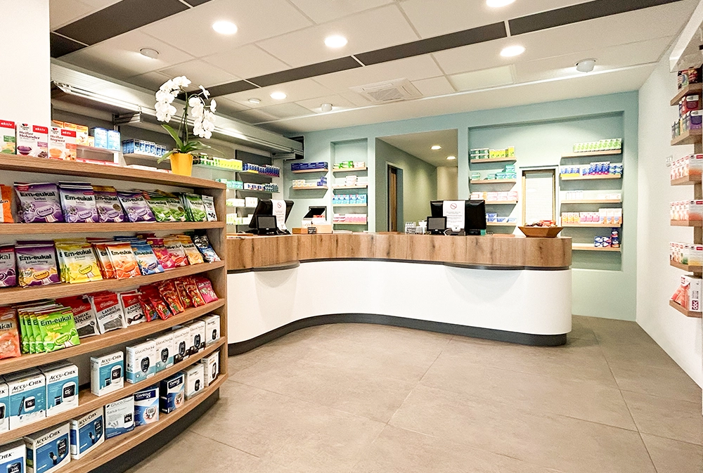 Innenansicht der Bären Apotheke Duisburg: Ein moderner Verkaufsraum mit geschwungenem Holztresen, gut beleuchtetem Kassenbereich und einem Regal mit Em-eukal Bonbons und Gesundheitsprodukten im Vordergrund. Die Rückwand ist mit Apothekenprodukten in farblich sortierten Regalen bestückt.
