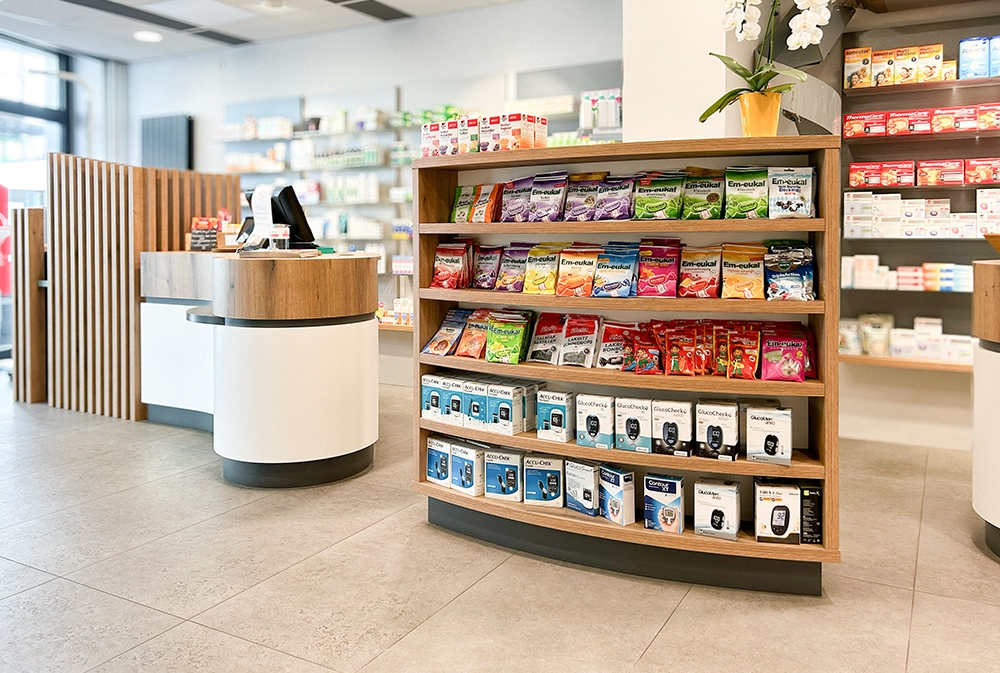 Innenaufnahme der Bären Apotheke Duisburg: Im Vordergrund ein gut sortiertes Regal mit Em-eukal Bonbons und Blutzuckermessgeräten. Dahinter der moderne, geschwungene Beratungstresen und Regale mit Gesundheitsprodukten in heller, freundlicher Umgebung.