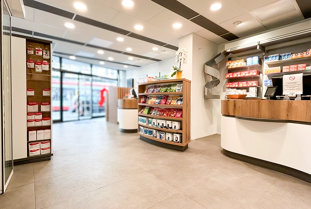 Innenaufnahme der Bären Apotheke Duisburg: Moderner Verkaufsraum mit hellen Böden, stilvoller Beleuchtung und gut sortierten Regalen mit Gesundheits- und Pflegeprodukten. Im Hintergrund ist der Kassenbereich zu sehen. Große Fensterfront mit Blick auf eine vorbeifahrende Straßenbahn.