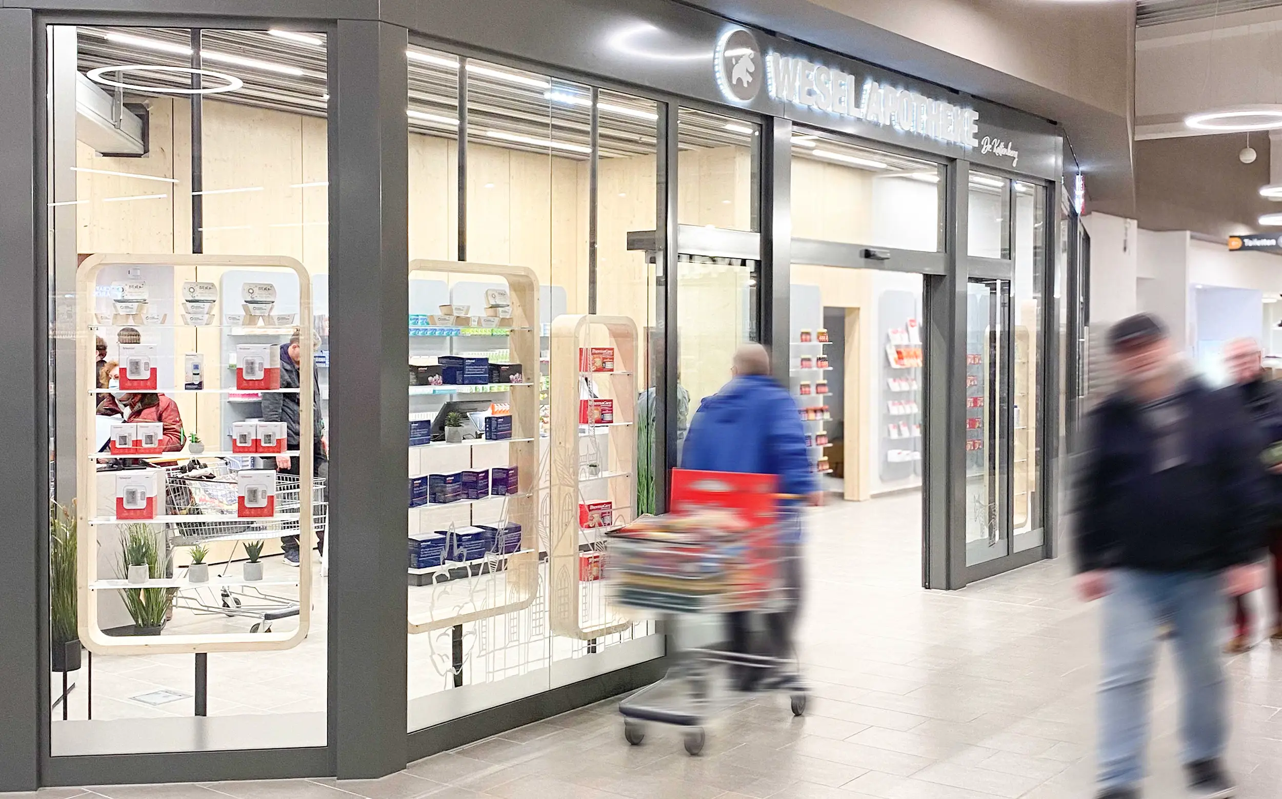 Die moderne Eingangsfassade der Wesel Apotheke im Einkaufszentrum mit großen Glasfenstern und hell beleuchtetem Innenraum. Im Vordergrund gehen mehrere Menschen mit Einkaufswagen vorbei.