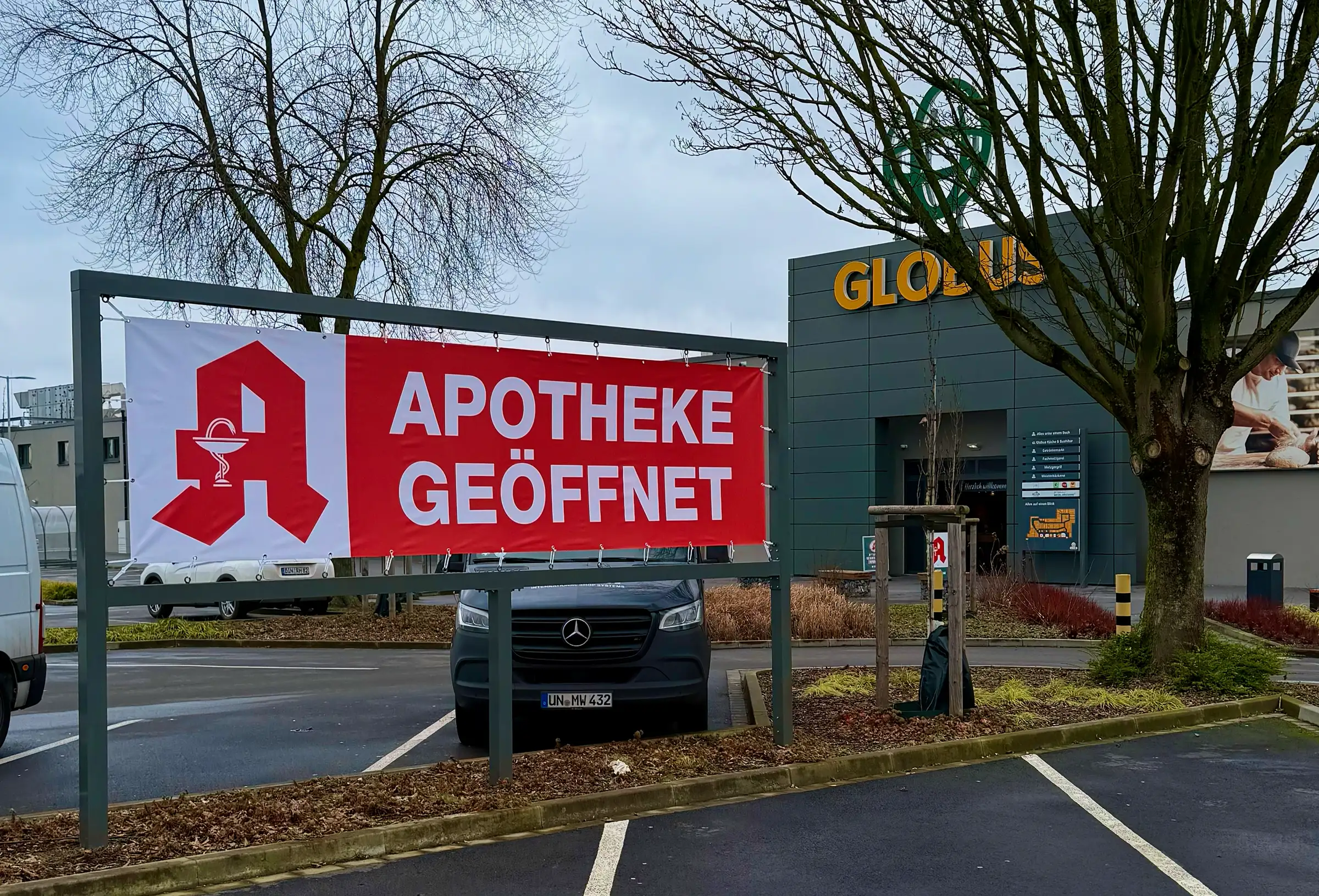 Ein großes rotes Banner mit der Aufschrift „Apotheke geöffnet“ hängt gut sichtbar auf einem Parkplatz vor einem Globus-Markt. Daneben ist das rote Apotheken-A zu sehen.